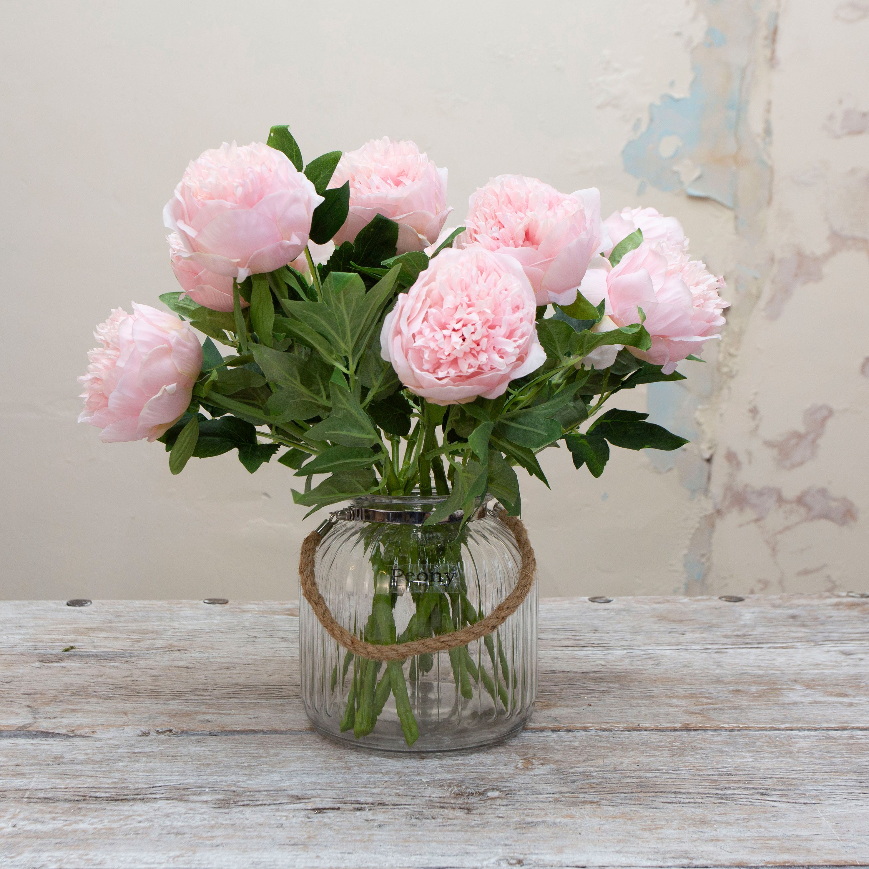 Artificial pale pink peony stems displayed in a glass vase, showcasing full, rounded blooms with lush green leaves