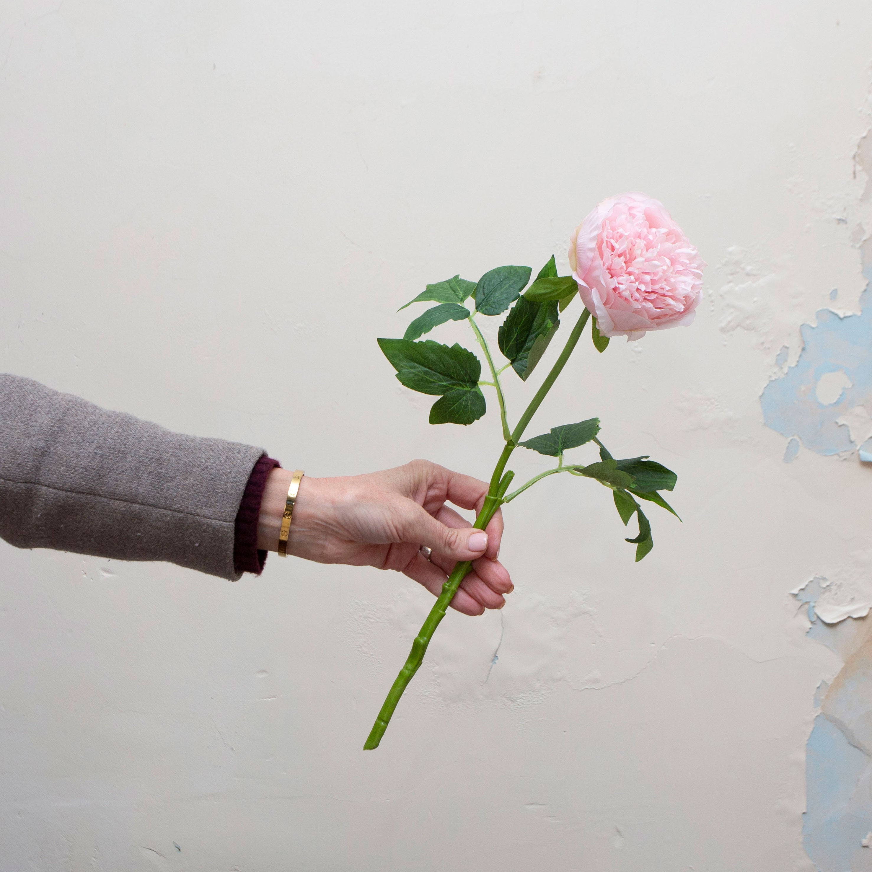 Artificial pale pink peony stem being held in hand, featuring a softly layered bloom with realistic green foliage