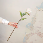 Artificial white peony stem held in hand, featuring a full ruffled bloom with subtle blush detailing and green leaves