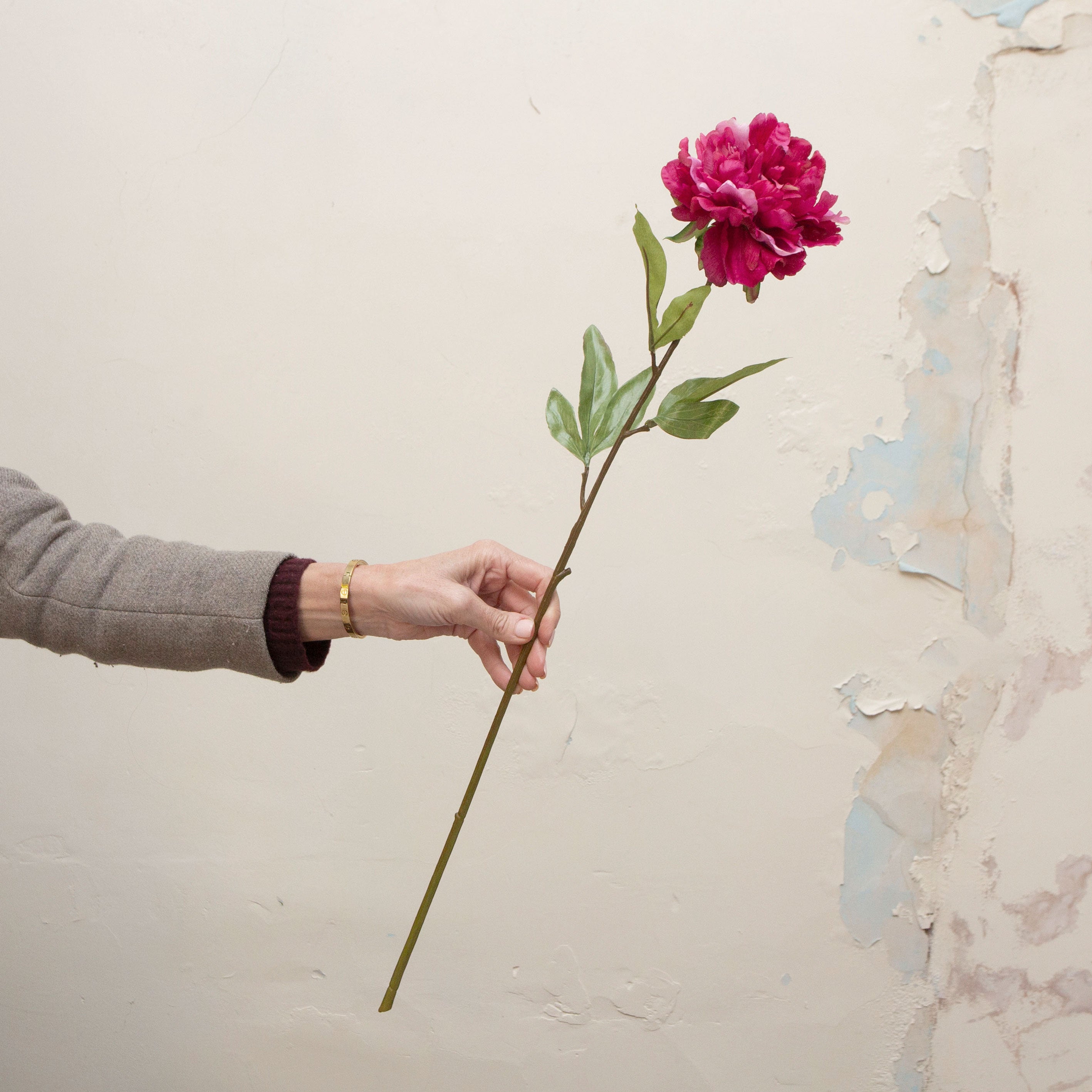 Artificial deep pink peony stem held in hand, featuring a full ruffled bloom and green leaves.