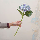 Artificial soft blue lilac hydrangea stem held in hand, featuring rounded flower clusters and green leaves on a sturdy stem