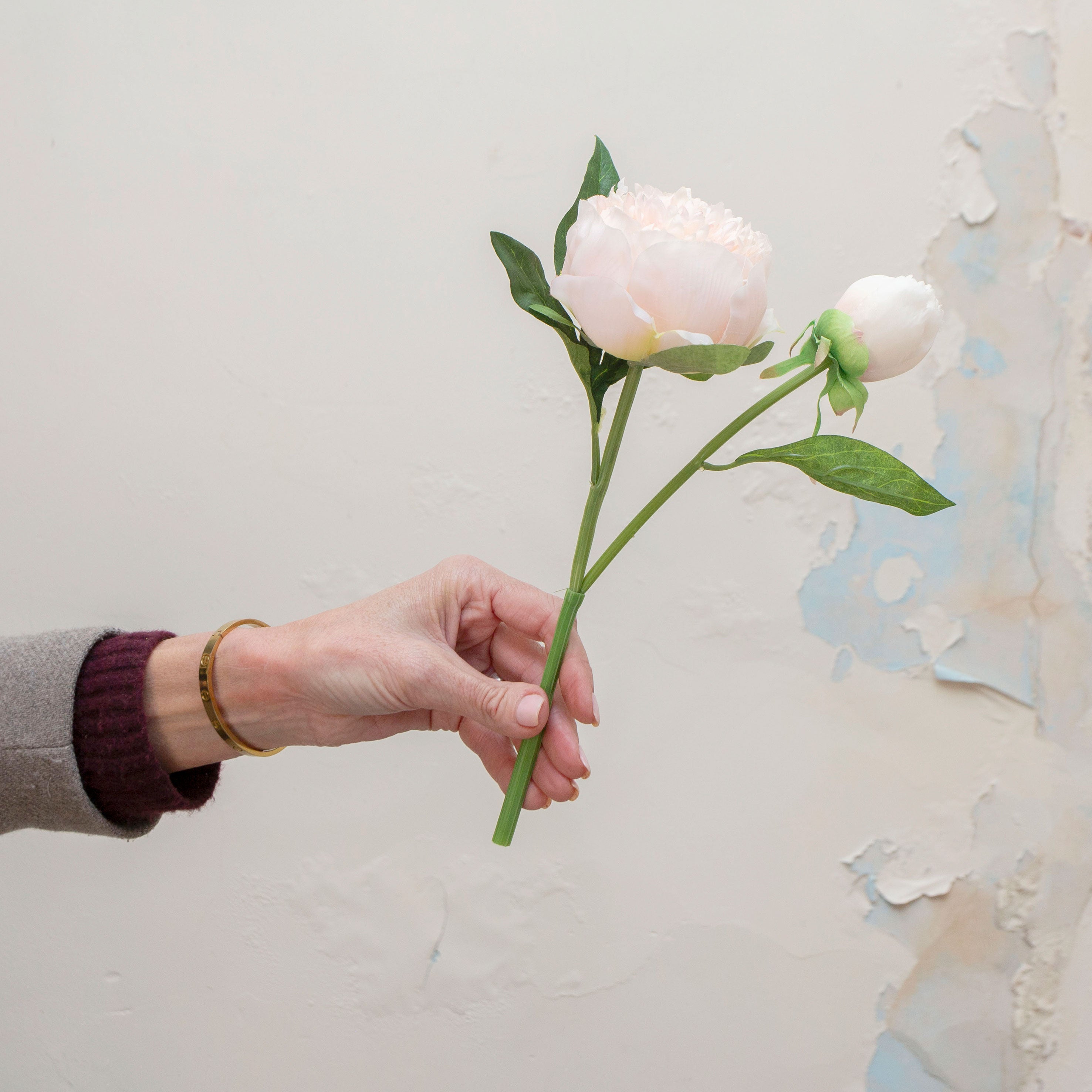 Artificial soft blush peach peony stem held in hand, featuring a full open bloom, closed buds, and green foliage.