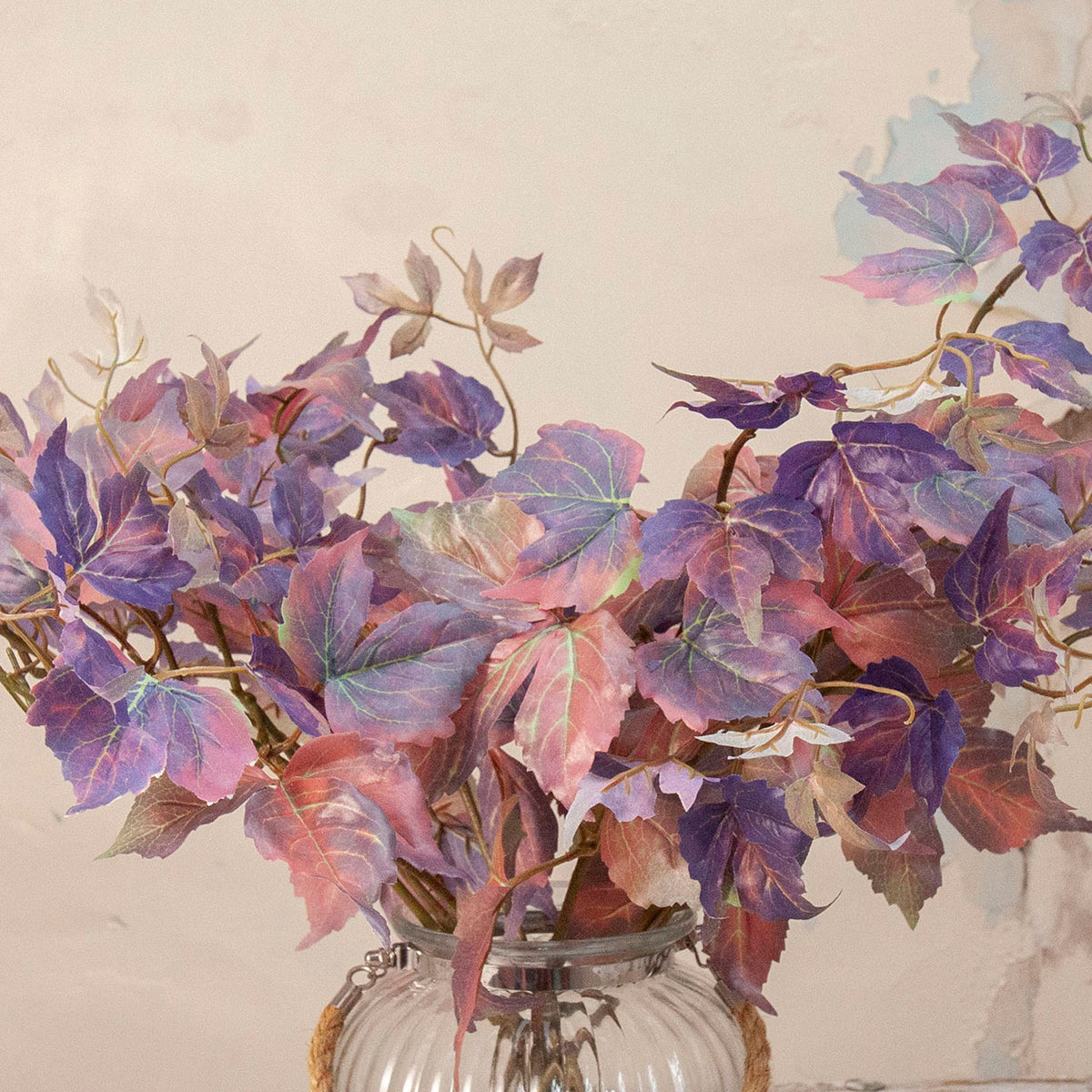 Close up of Artificial Leaf Foliage Stems with trailing purple, burgundy and muted autumn-toned leaves displayed in a clear ribbed glass vase
