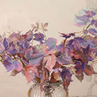 Close up of Artificial Leaf Foliage Stems with trailing purple, burgundy and muted autumn-toned leaves displayed in a clear ribbed glass vase