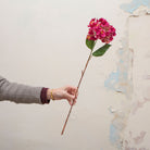 Artificial bright pink hydrangea stem held in hand, featuring clustered blooms and green leaves on a woody stem.