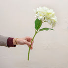 Artificial white hydrangea single stem being held in hand, featuring a full rounded flower head with soft ivory-white petals and realistic green foliage