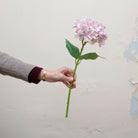 Artificial lilac-pink hydrangea single stem being held in hand, featuring a rounded cluster of soft pastel petals with subtle lilac and blush tones, paired with realistic green leaves on a sturdy stem