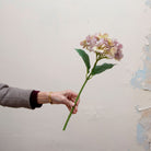 Artificial hydrangea stem being held in hand, featuring a rounded flower head in soft lilac and blush tones with subtle green undertones and realistic textured leaves