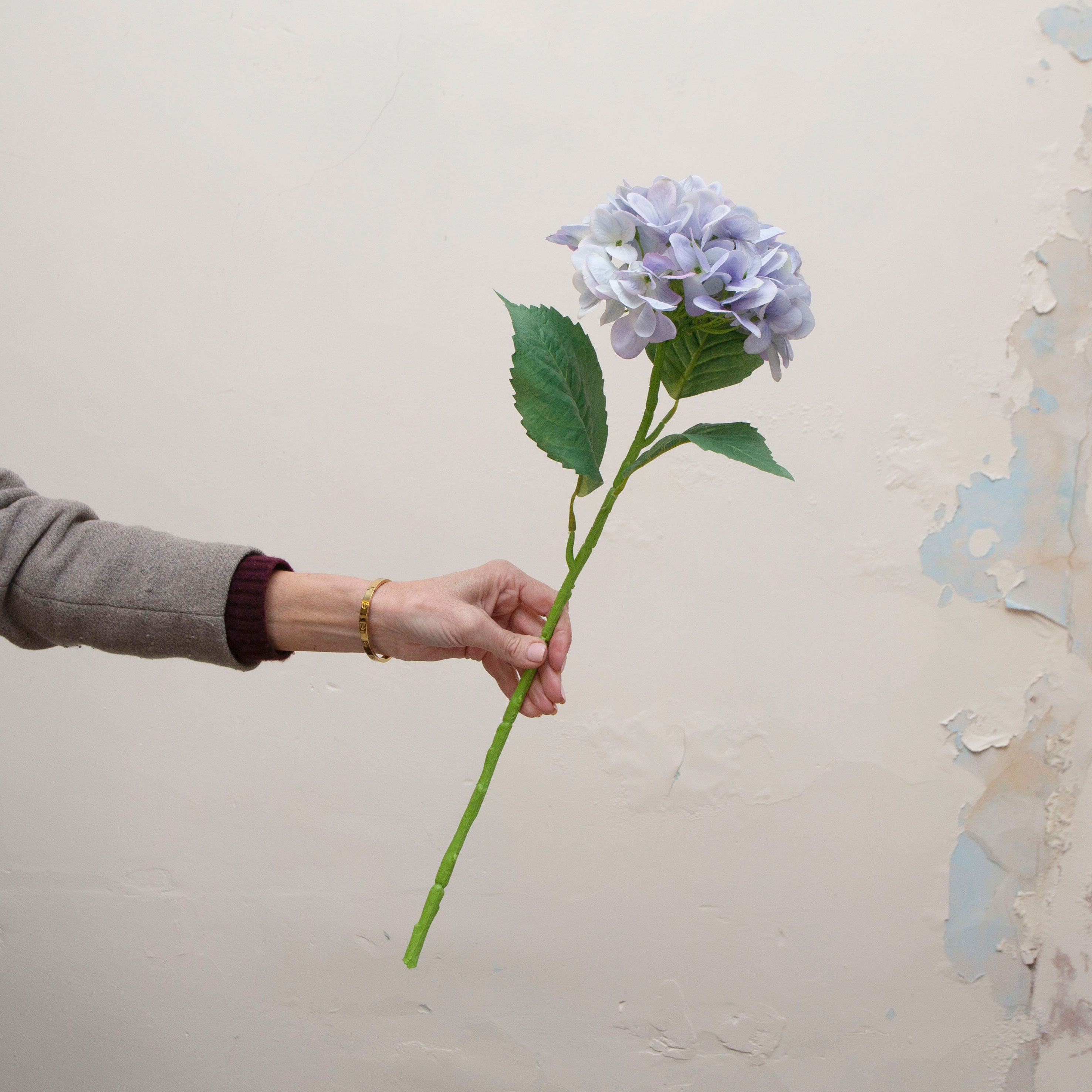 Artificial pale blue hydrangea stem being held in hand, featuring a full rounded hydrangea flower head with soft pastel blue petals and realistic green foliage on a sturdy stem
