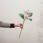 Artificial lilac hydrangea stem being held in hand, featuring a full rounded flower head with soft lilac and pale lavender petals and realistic green leaves on a sturdy stem