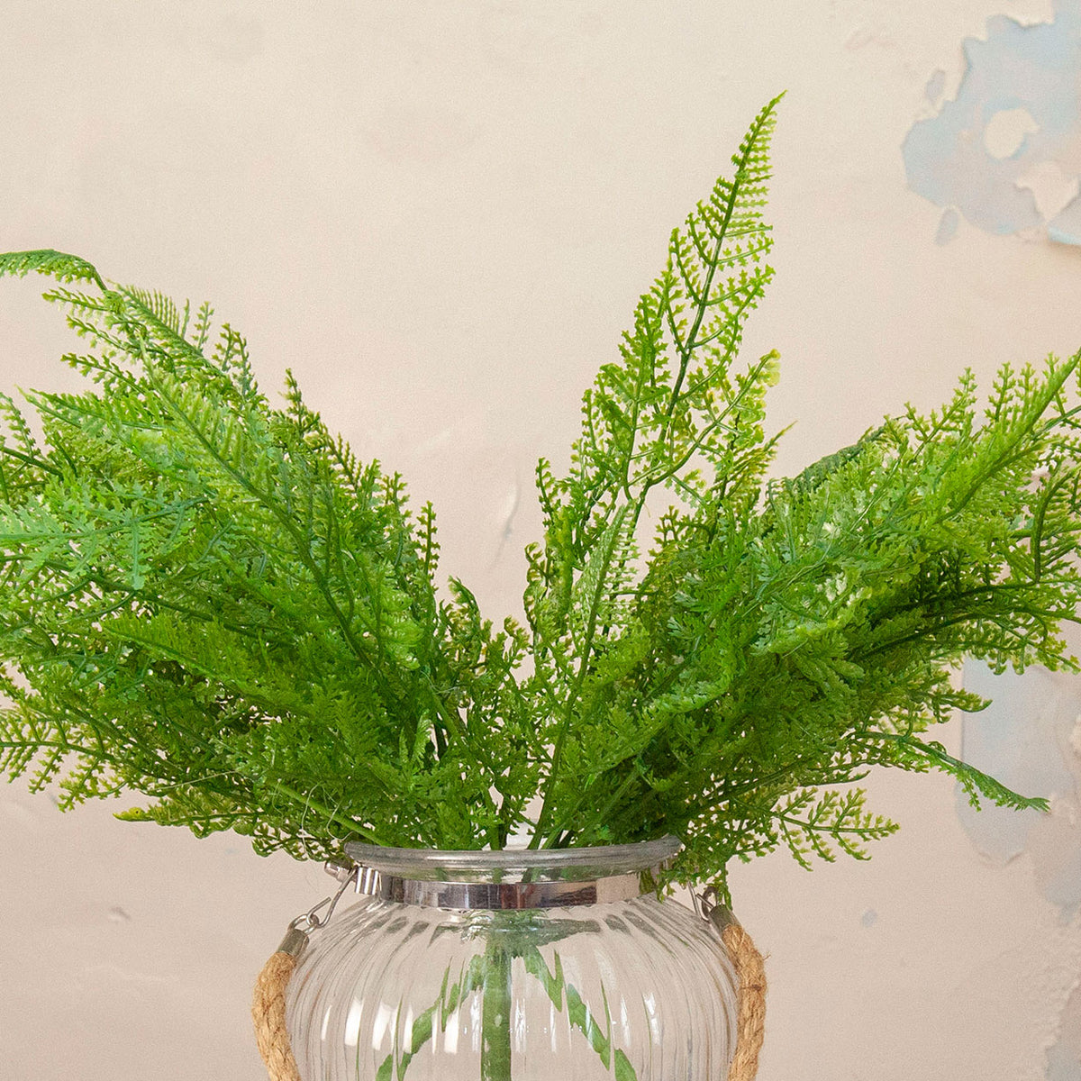 Close up of Artificial Fern Bush Stems with lush, feathery green fern foliage displayed in a clear ribbed glass vase with rope handle