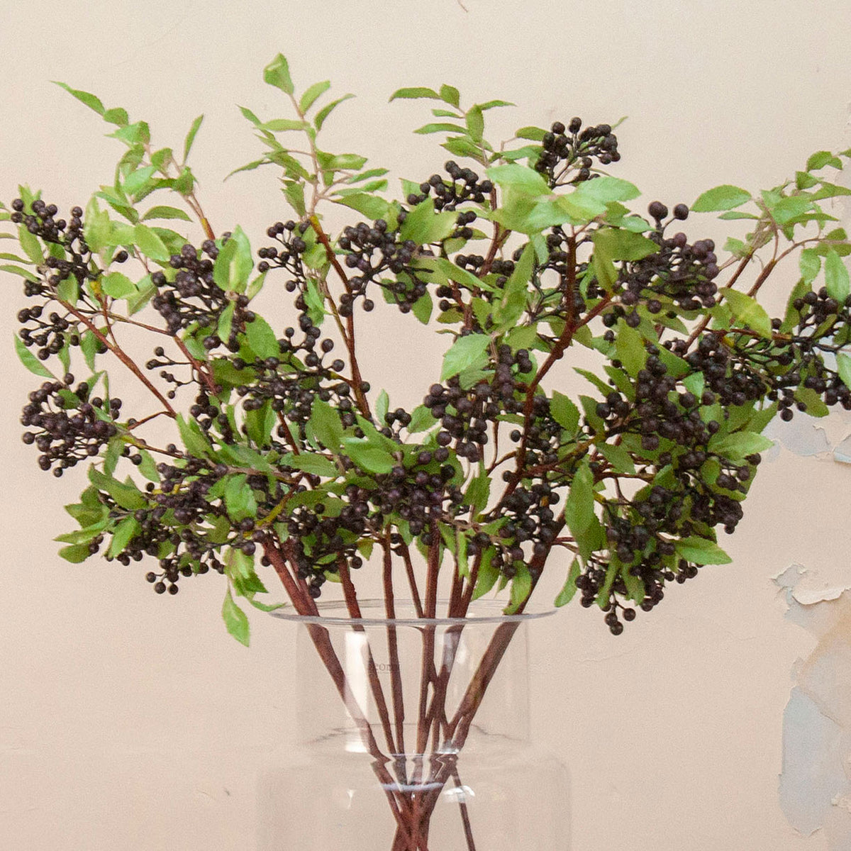 Close up Artificial Berry Stems with clusters of dark purple-black berries, fresh green leaves and natural brown branches displayed in a clear glass vase