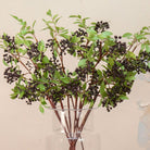 Close up Artificial Berry Stems with clusters of dark purple-black berries, fresh green leaves and natural brown branches displayed in a clear glass vase