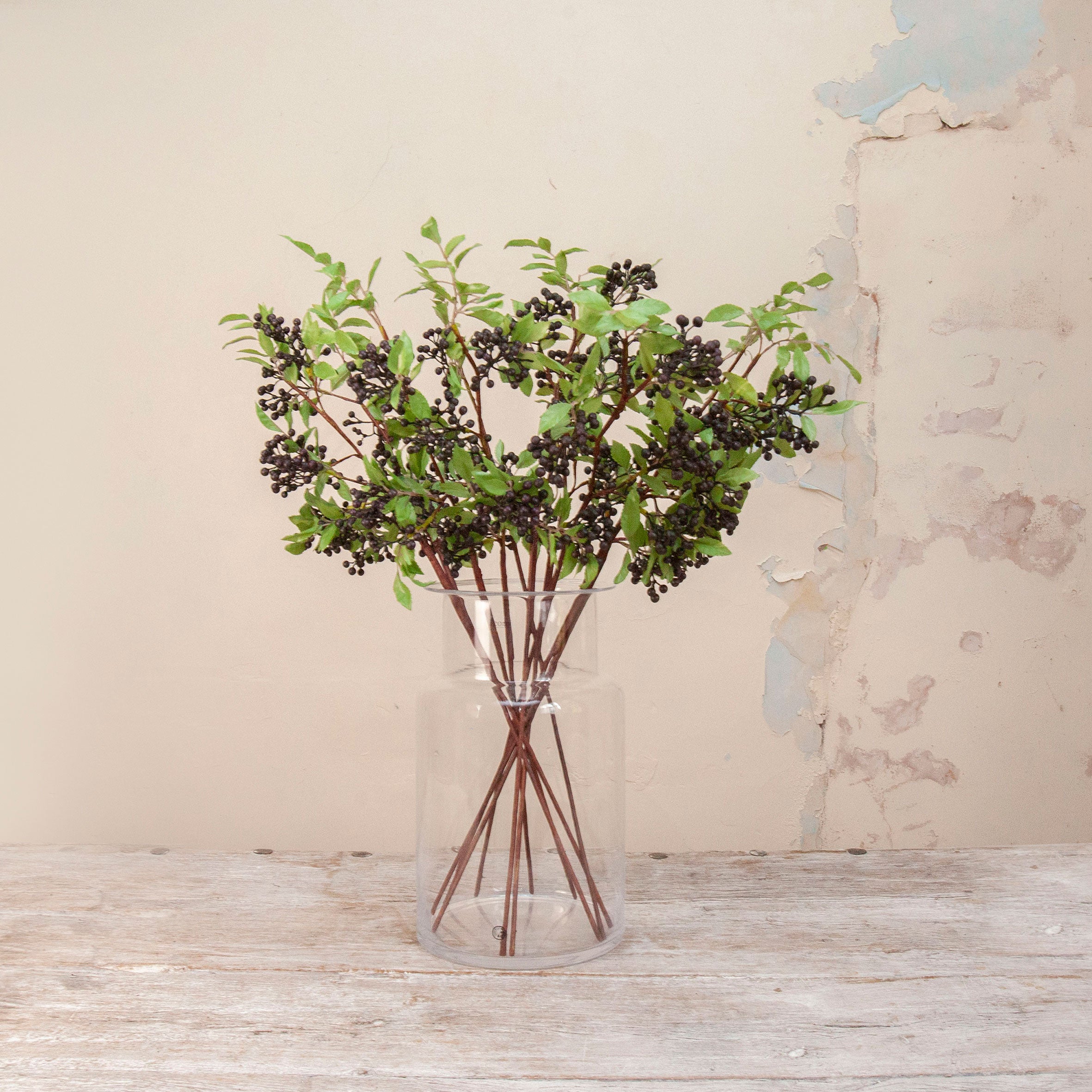 Artificial Berry Stems with clusters of dark purple-black berries, fresh green leaves and natural brown branches displayed in a clear glass vase
