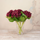 Artificial Hydrangea Stems with rich burgundy hydrangea flower heads and realistic green leaves displayed in a clear glass vase