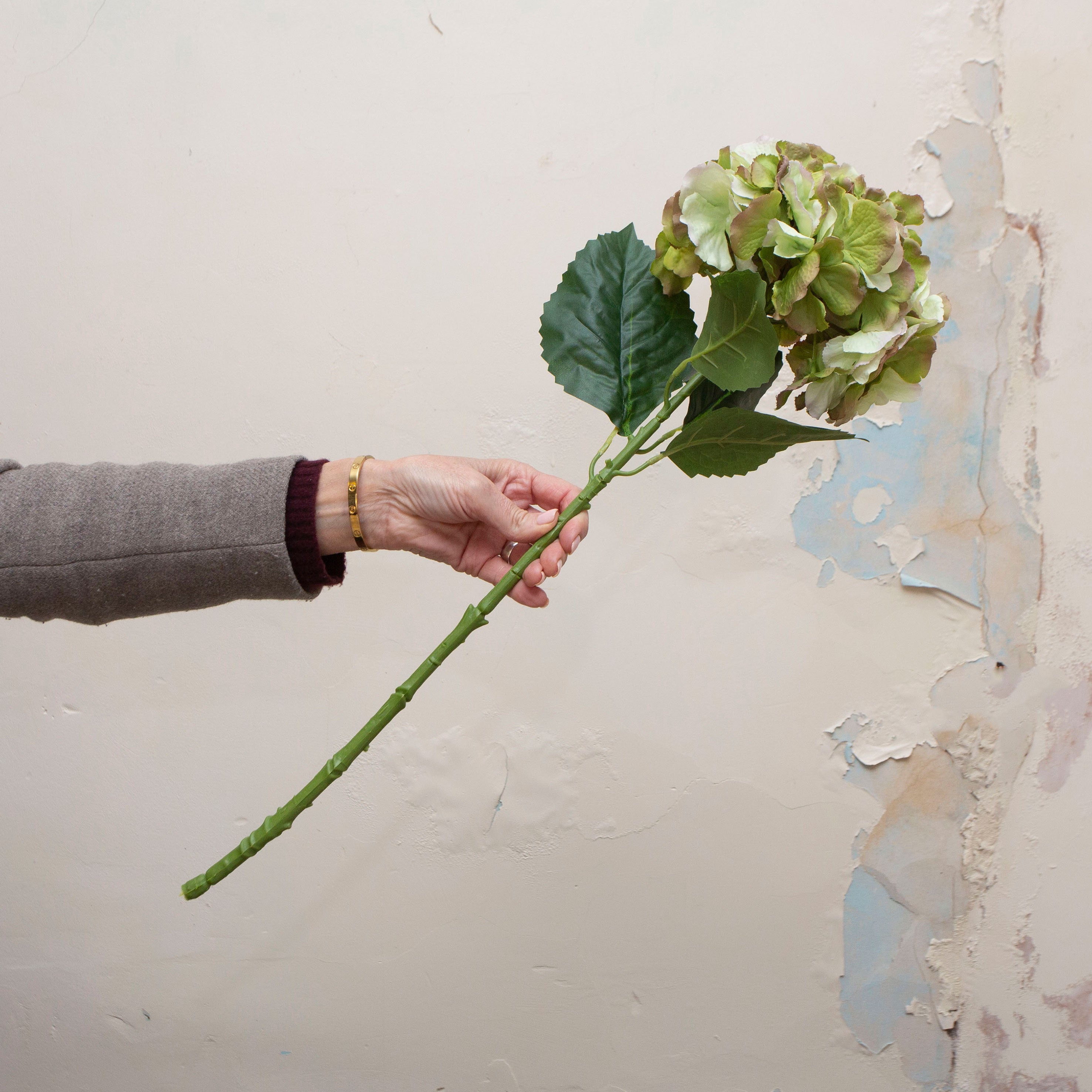 Artificial green hydrangea stem being held in hand, featuring a rounded hydrangea flower head with soft green petals and subtle blush edging, paired with realistic foliage on a sturdy stem