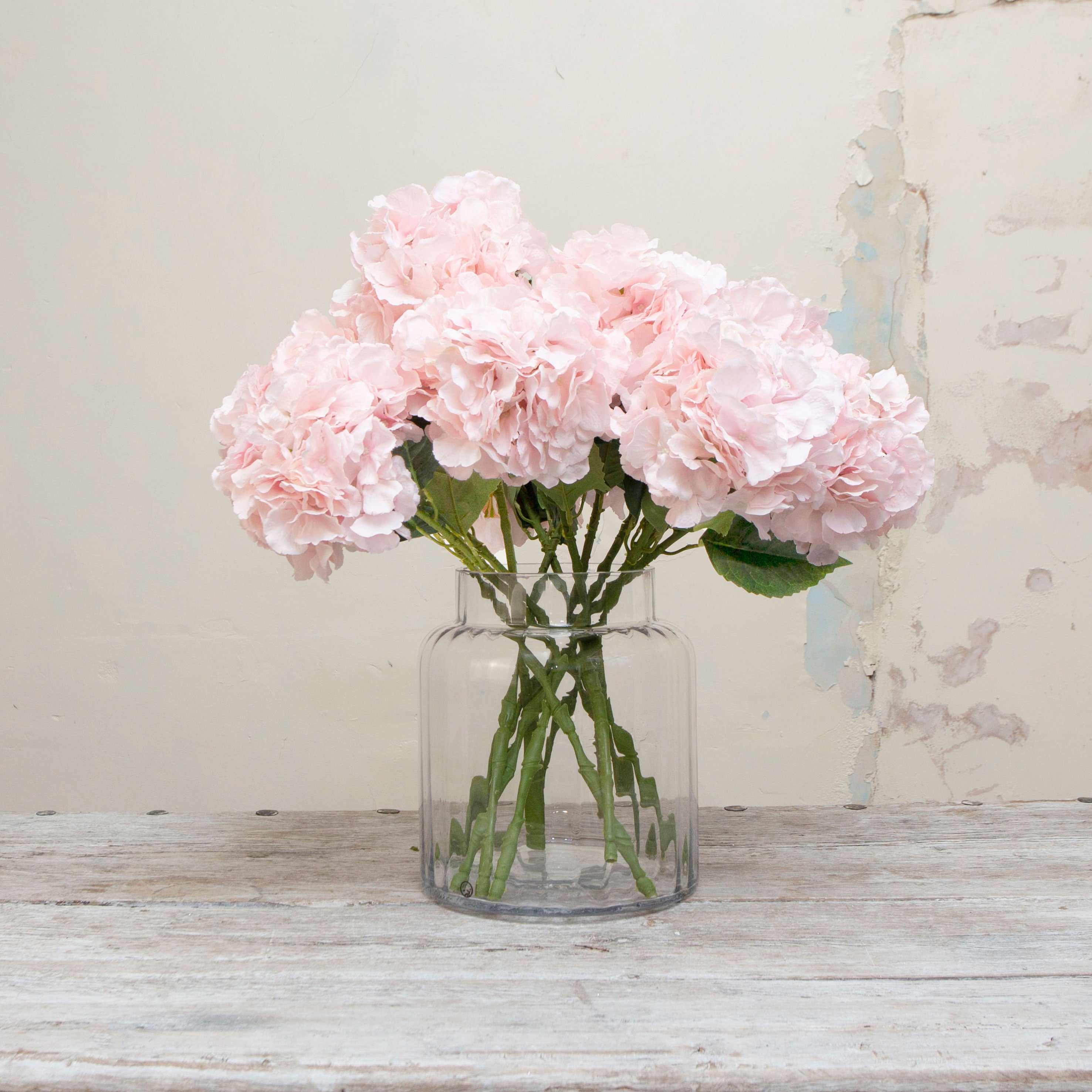 Artificial pale pink hydrangea stem displayed in a vase, showcasing a lush hydrangea bloom in soft blush-pink tones with natural-looking leaves and gentle colour variation