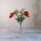 Artificial red poppy and patrinia arrangement with green foliage in a clear glass milk bottle vase.