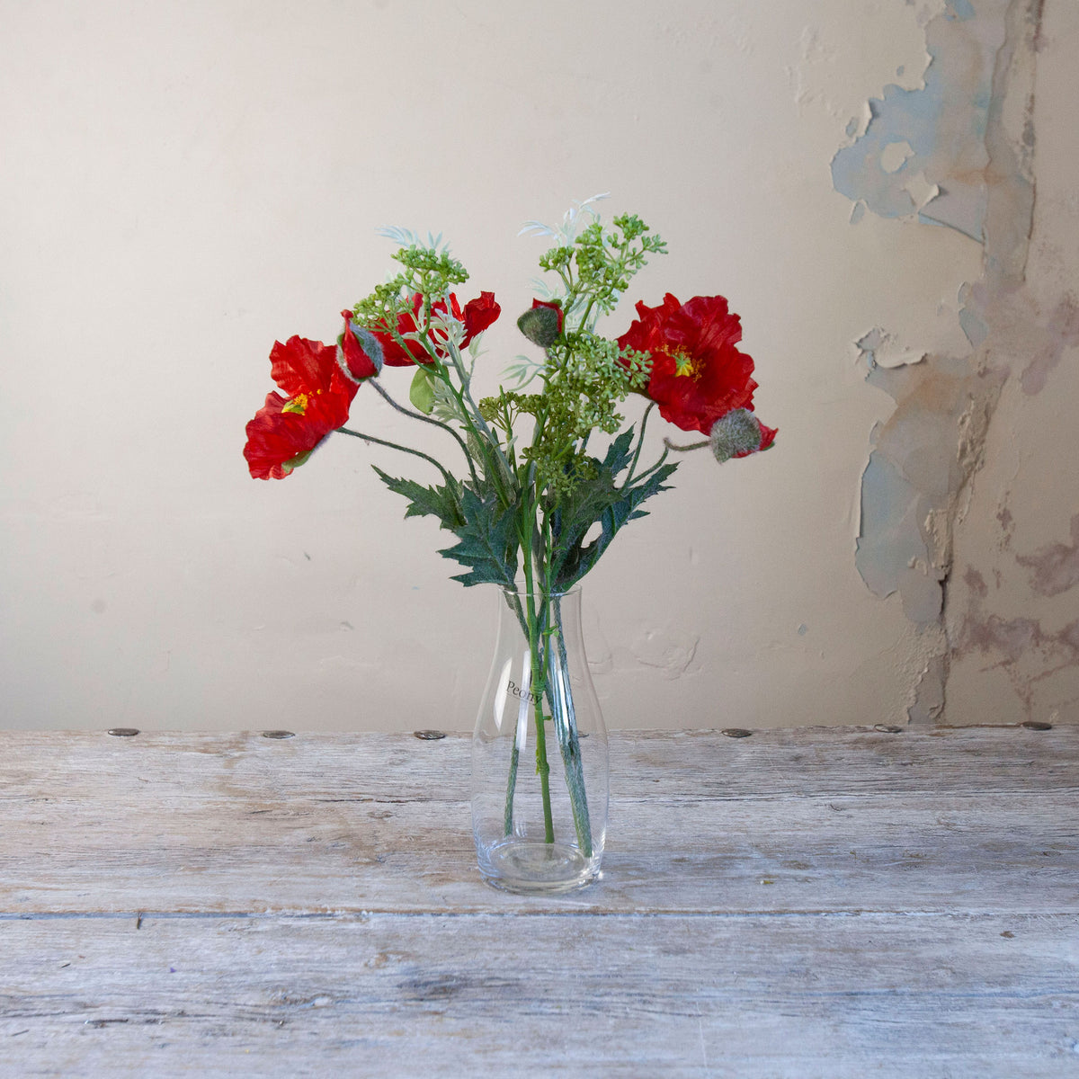 Artificial red poppy and patrinia arrangement with green foliage in a clear glass milk bottle vase.