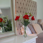 Artificial red poppy and patrinia arrangement with green foliage in a clear glass milk bottle vase, styled on a bedroom dressing table.