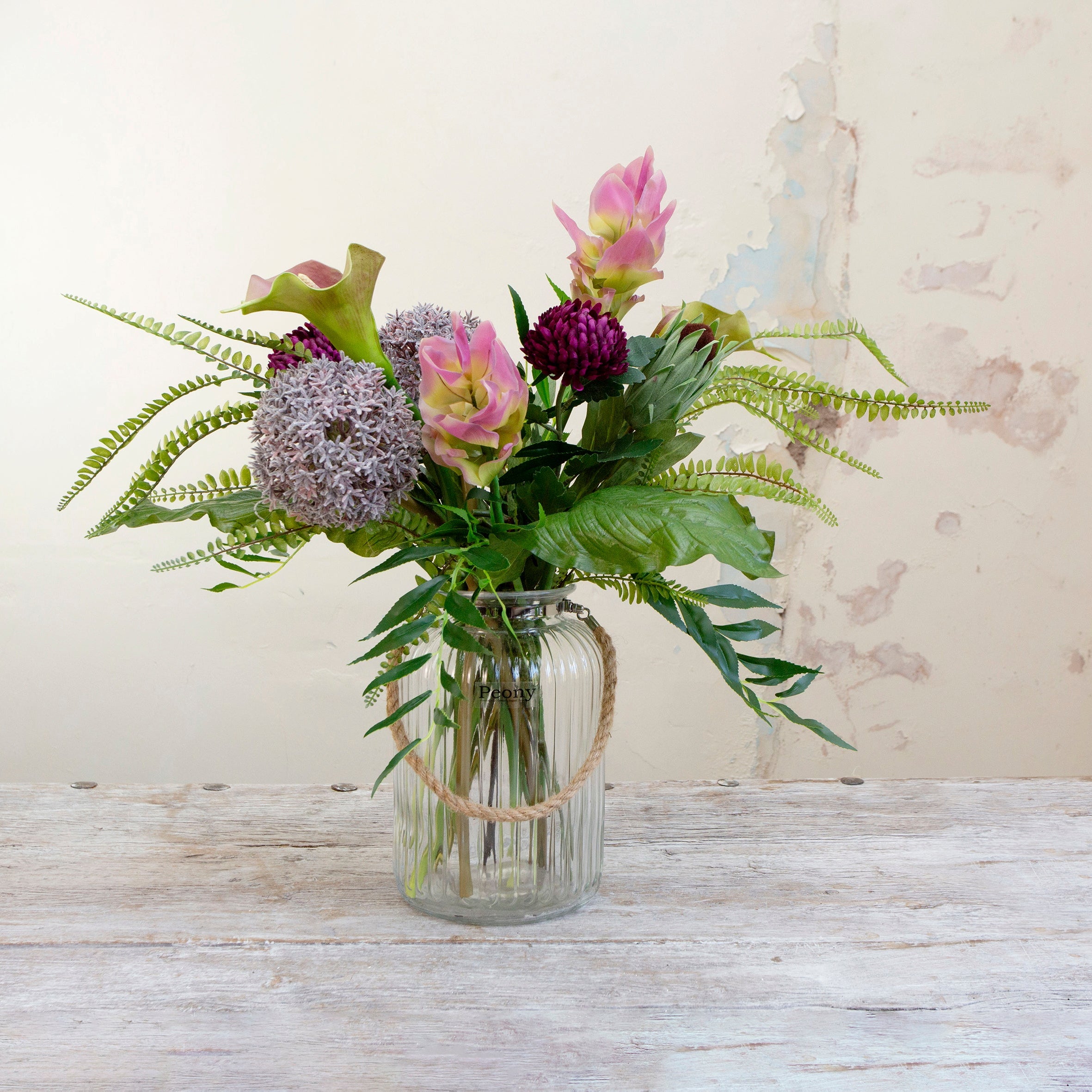 artificial bouquet with curcuma, alliums, protea, calla lilies, and fern foliage arranged in a clear ribbed glass lantern vase with rustic rope handles.