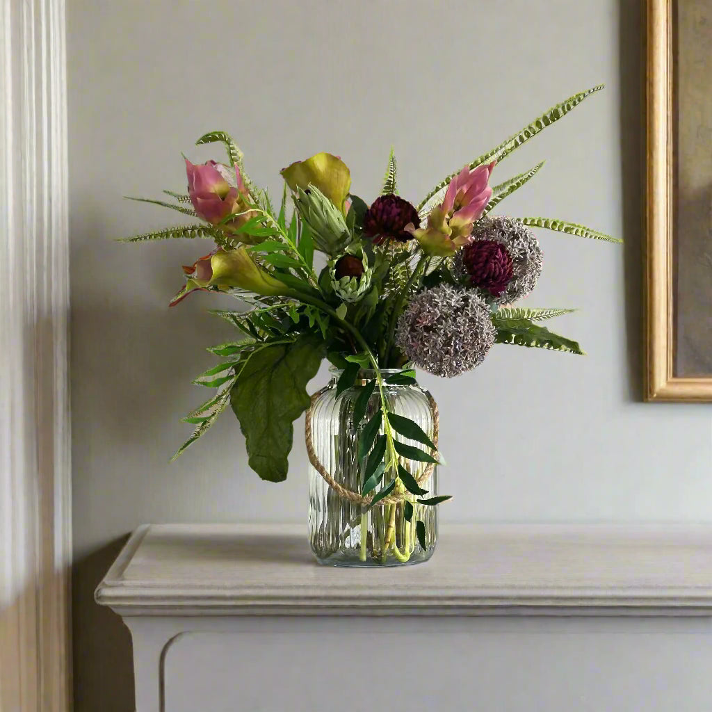 artificial bouquet with curcuma, alliums, protea, calla lilies, and fern foliage arranged in a clear ribbed glass lantern vase with rustic rope handles.