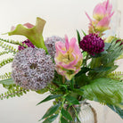 artificial bouquet with curcuma, alliums, protea, calla lilies, and fern foliage arranged in a clear ribbed glass lantern vase with rustic rope handles.