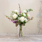 Artificial cream roses and lilac foxgloves arranged in a clear Grecian style clear glass vase