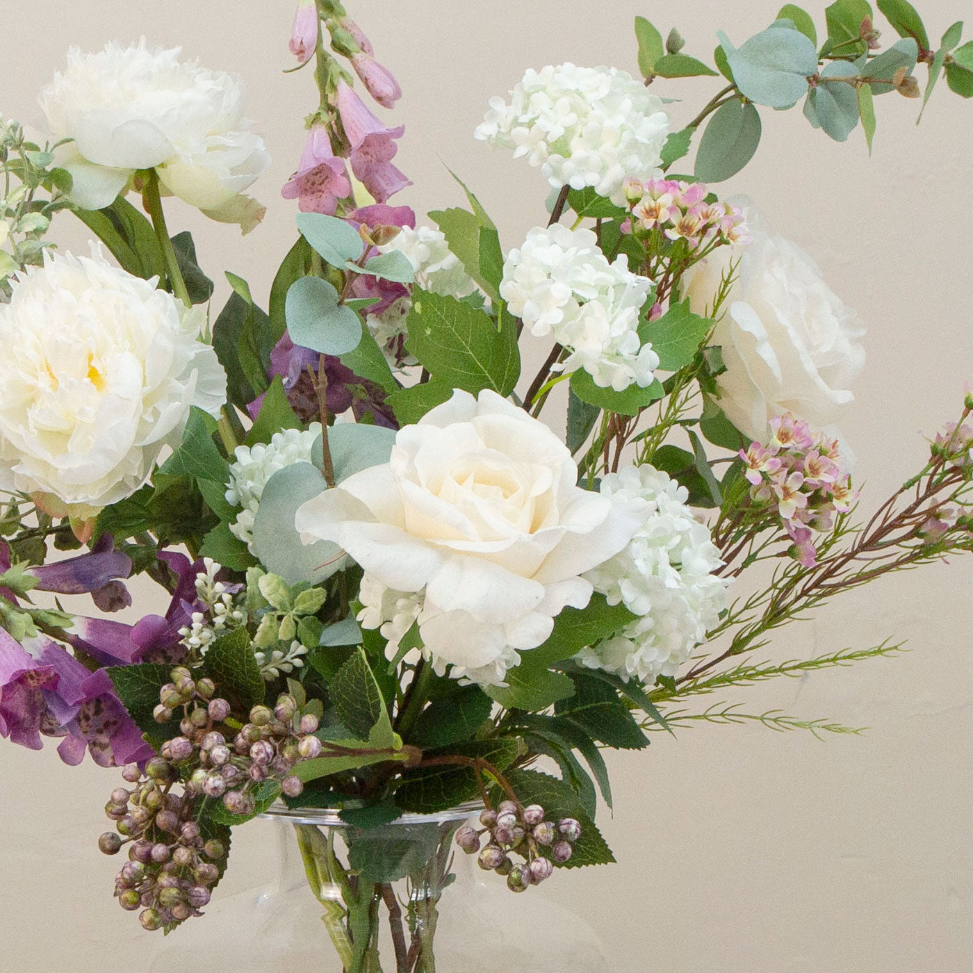Artificial cream roses and lilac foxgloves arranged in a clear Grecian style clear glass vase