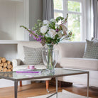 Artificial cream roses and lilac foxgloves arranged in a clear Grecian style clear glass vase
