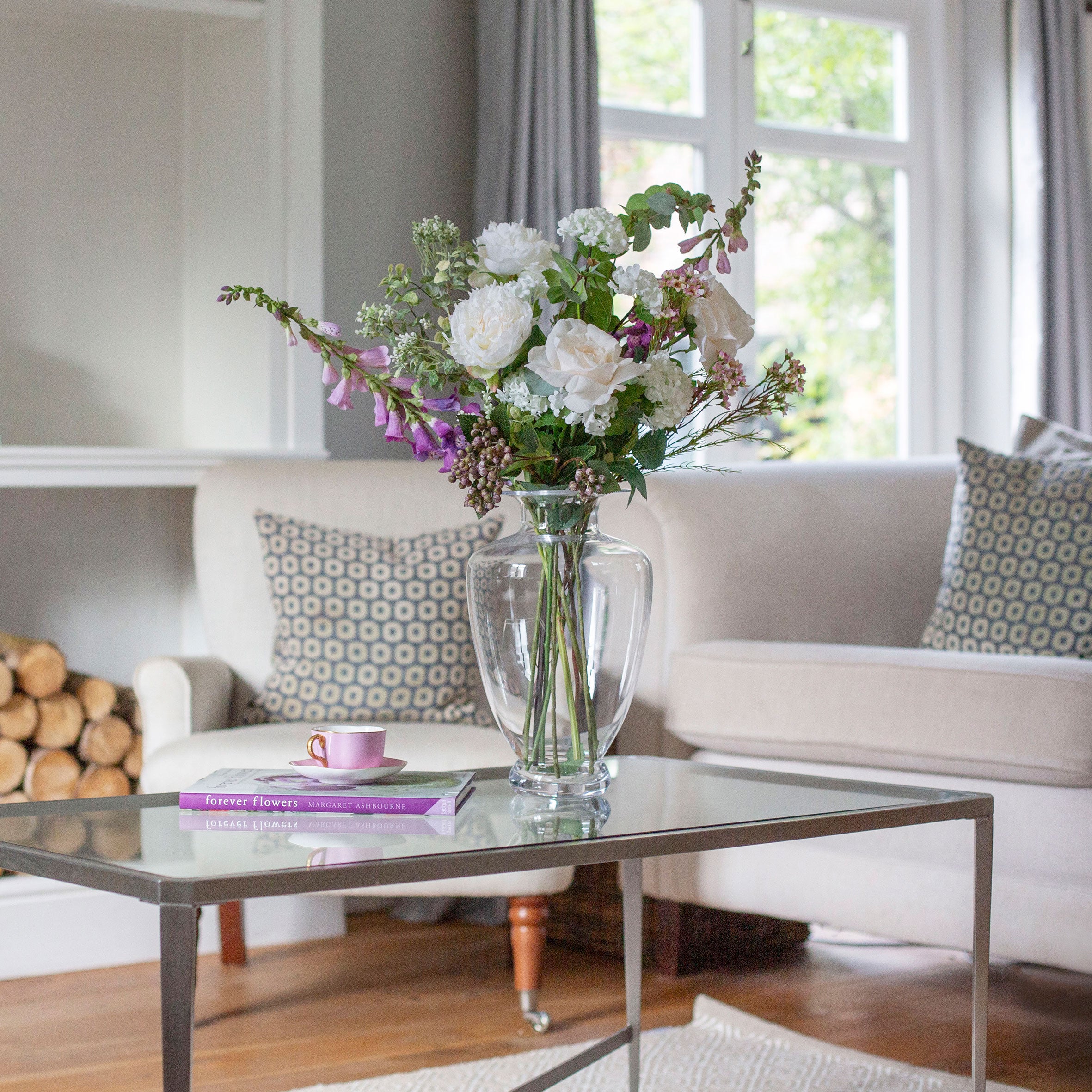Artificial cream roses and lilac foxgloves arranged in a clear Grecian style clear glass vase