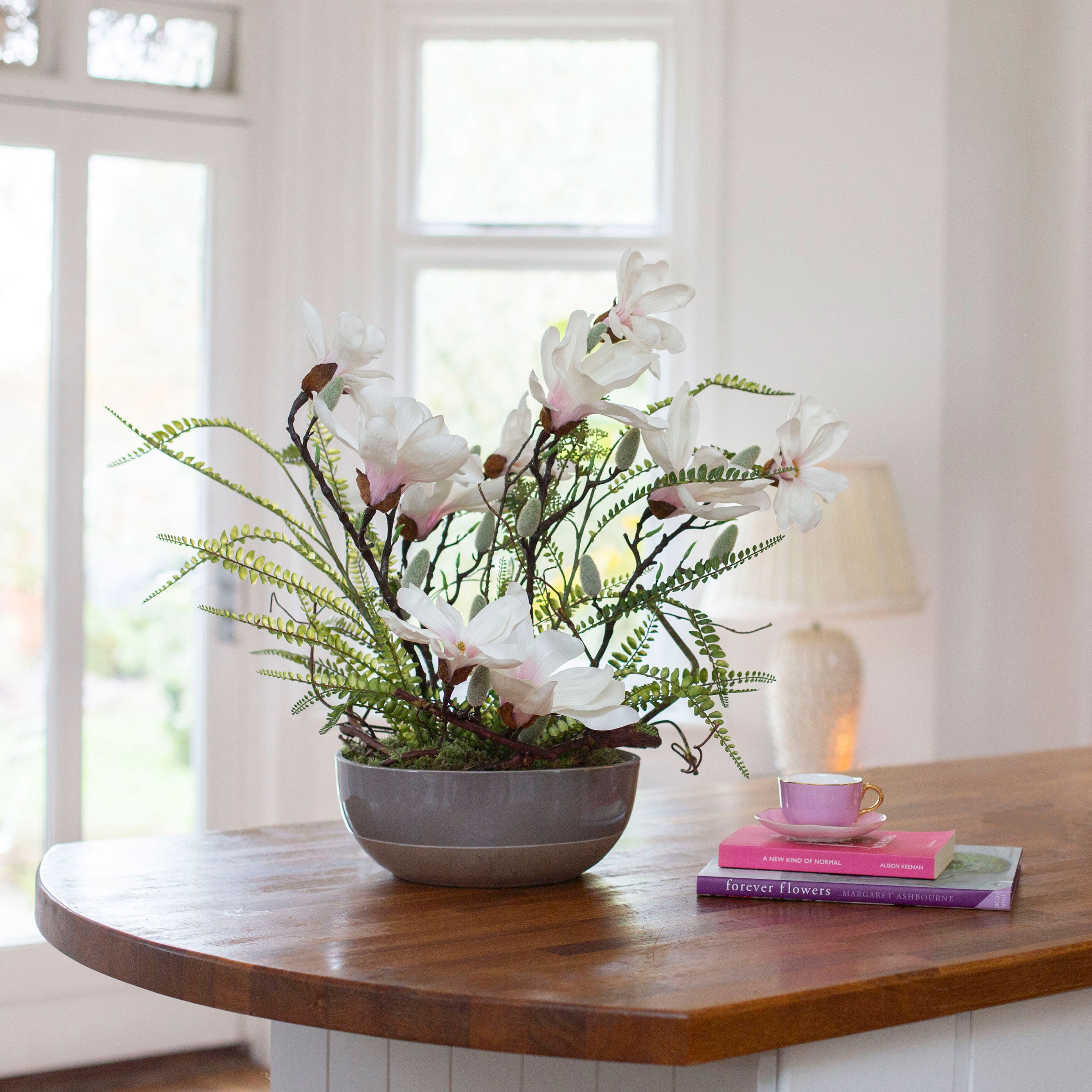 Artificial magnolia flowers and ferns arranged in a low grey banded bowl