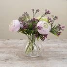 Artificial ranunculus and foliage arrangement in a clear glass garlic vase