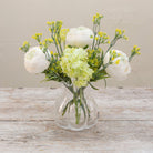 Artificial ranunculus and foliage arrangement in a clear glass garlic vase