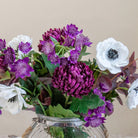 Close up of artificial chrysanthemum, anemone and astrantia bouquet in rich purple and white tones, displayed in a clear glass rope vase.