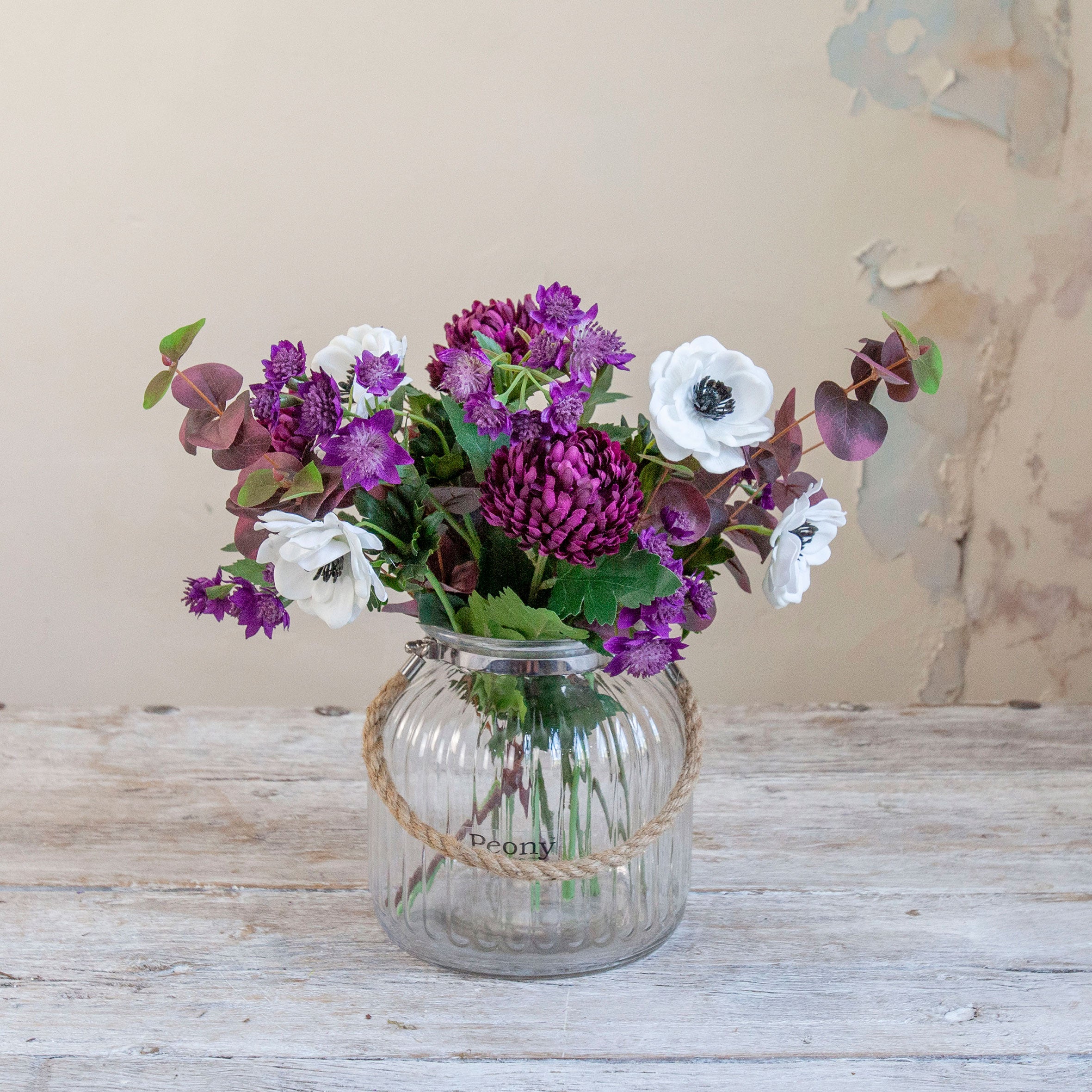 Artificial chrysanthemum, anemone and astrantia bouquet in rich purple and white tones, displayed in a clear glass rope vase.