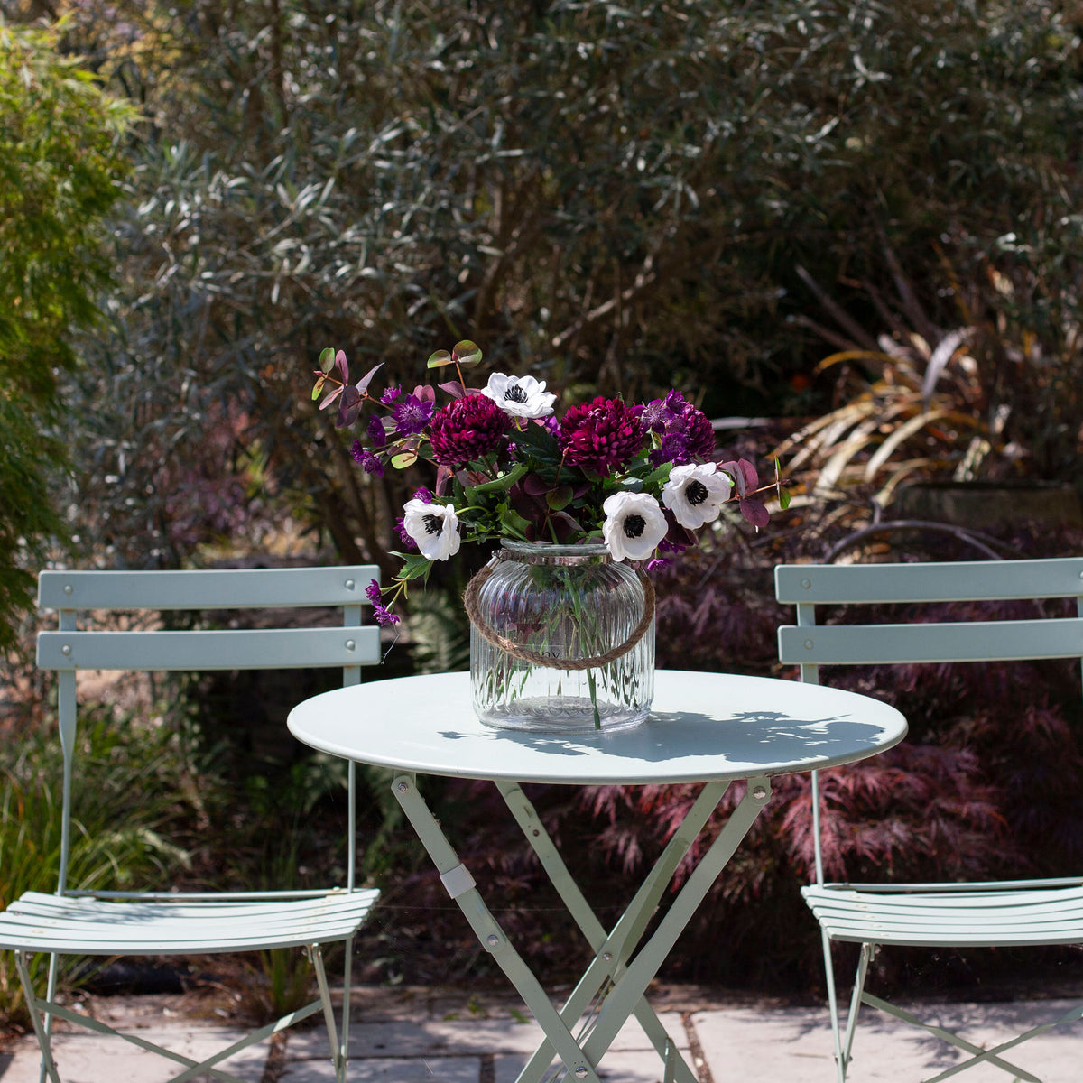 Artificial chrysanthemum, anemone and astrantia bouquet in rich purple and white tones, displayed in a clear glass rope vase on an outdoor garden table.