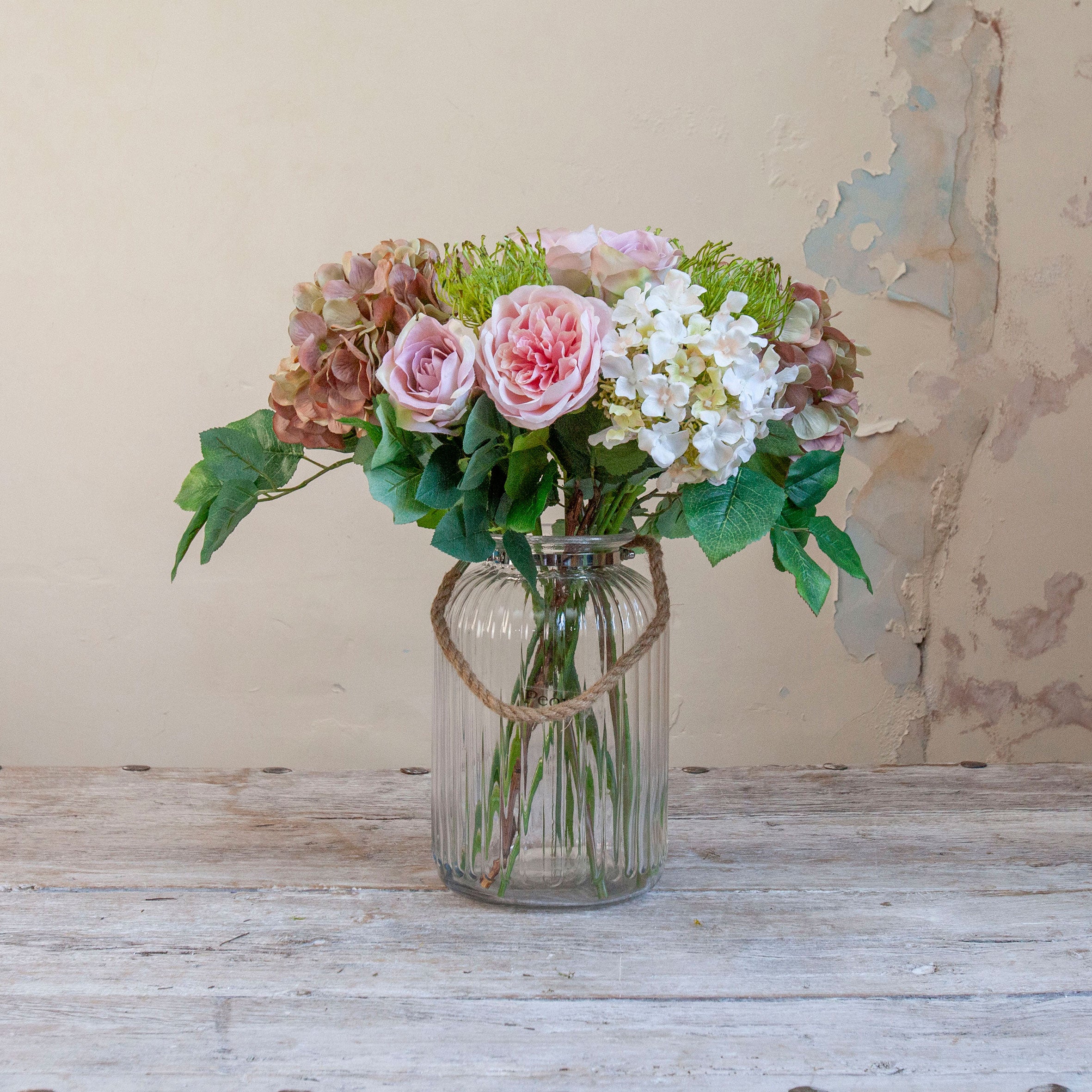 Artificial hydrangea, rose and chrysanthemum bouquet in soft pink, white and muted autumn tones, displayed in a clear glass rope vase.