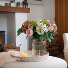 Artificial hydrangea, rose and chrysanthemum bouquet in soft pink, white and muted autumn tones, displayed in a clear glass rope vase in a cosy living room.