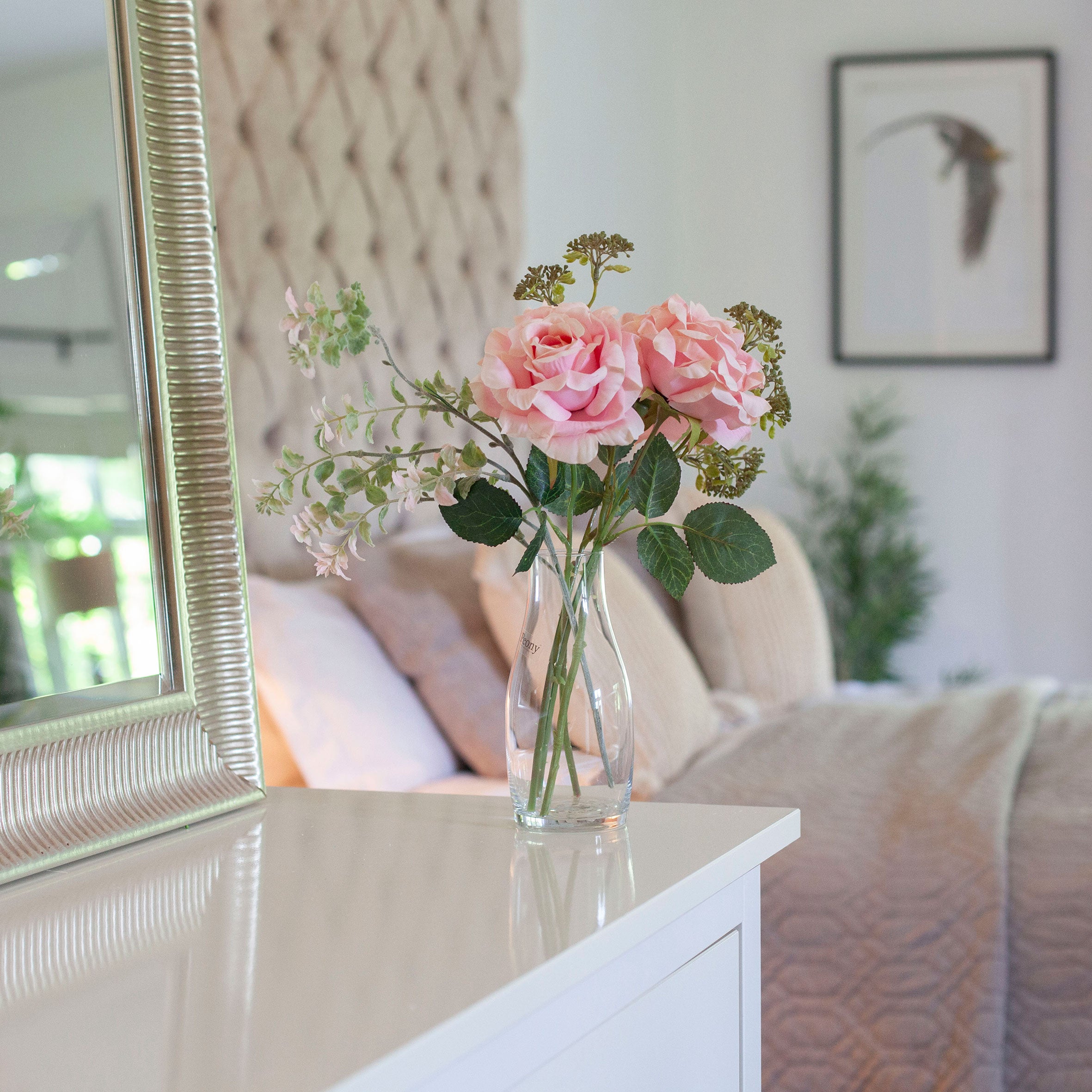Artificial pink roses and patrinia set in a clear glass milk bottle vase, styled on a bedroom dressing table.