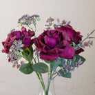 Close-up of artificial burgundy roses and patrinia in a clear glass milk bottle vase.