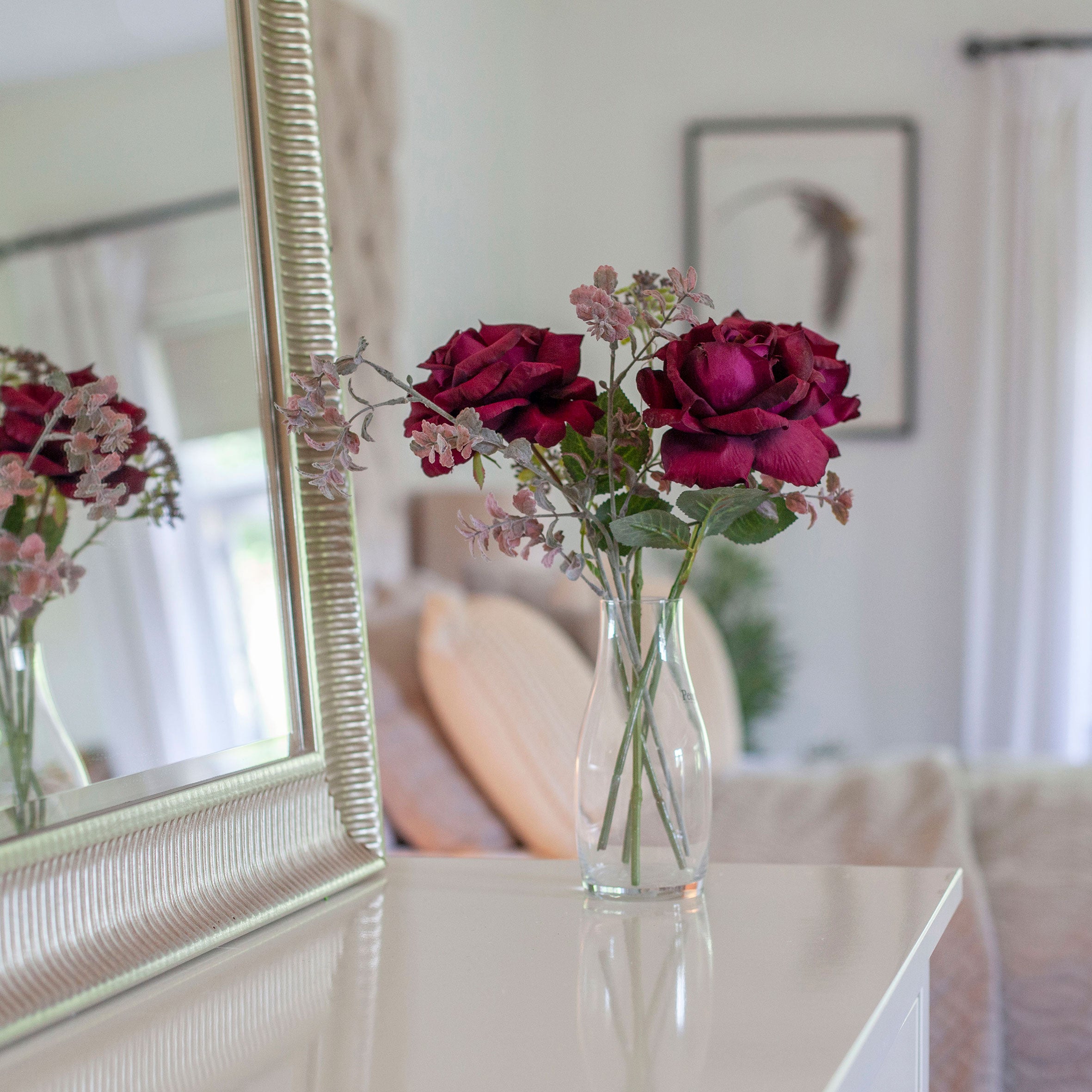 Artificial white roses and purple/burgundy set in a clear glass milk bottle vase, styled on a bedroom dressing table.
