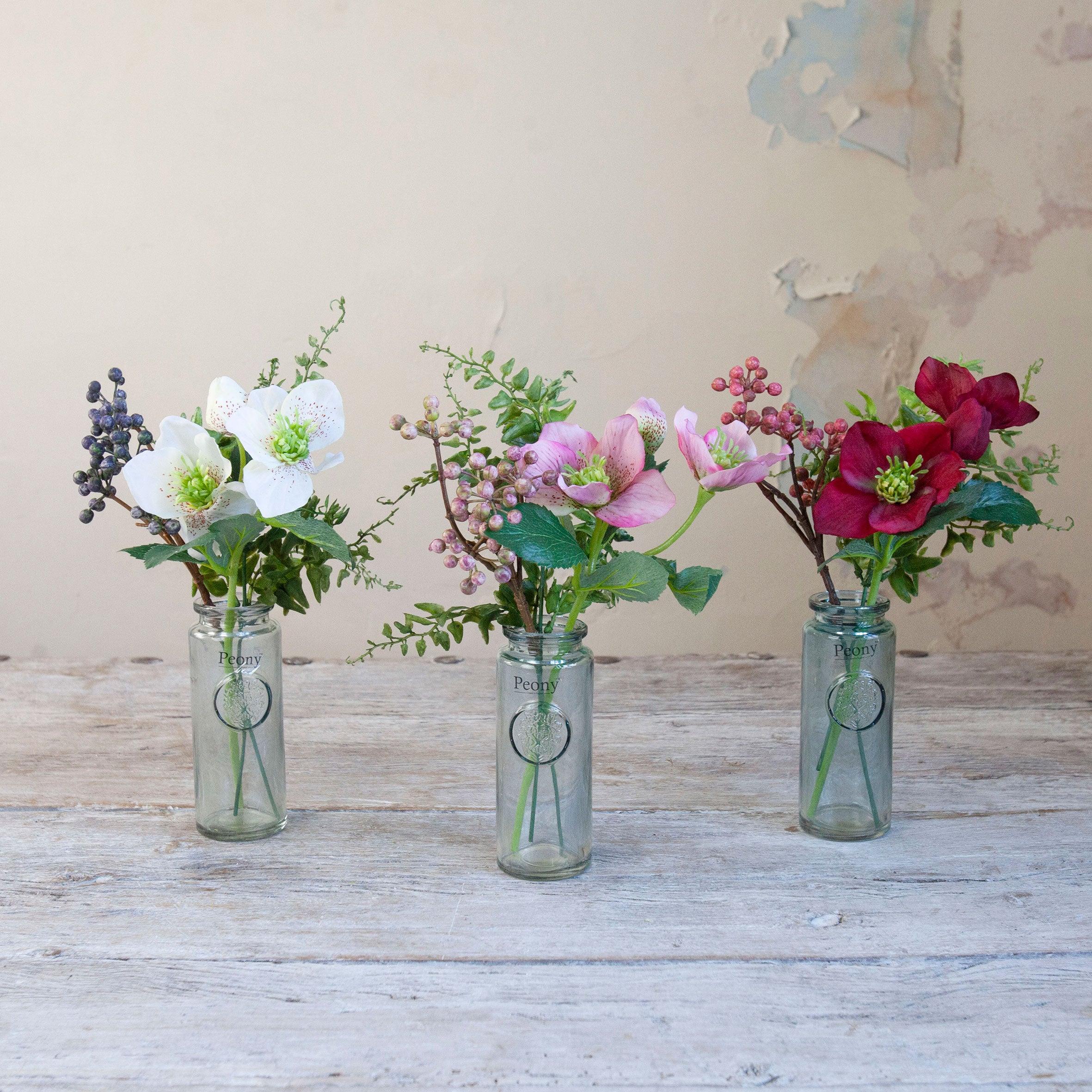 Set of three artificial hellebore and berry arrangements in white, pink and burgundy, arranged in individual glass bottle vases.