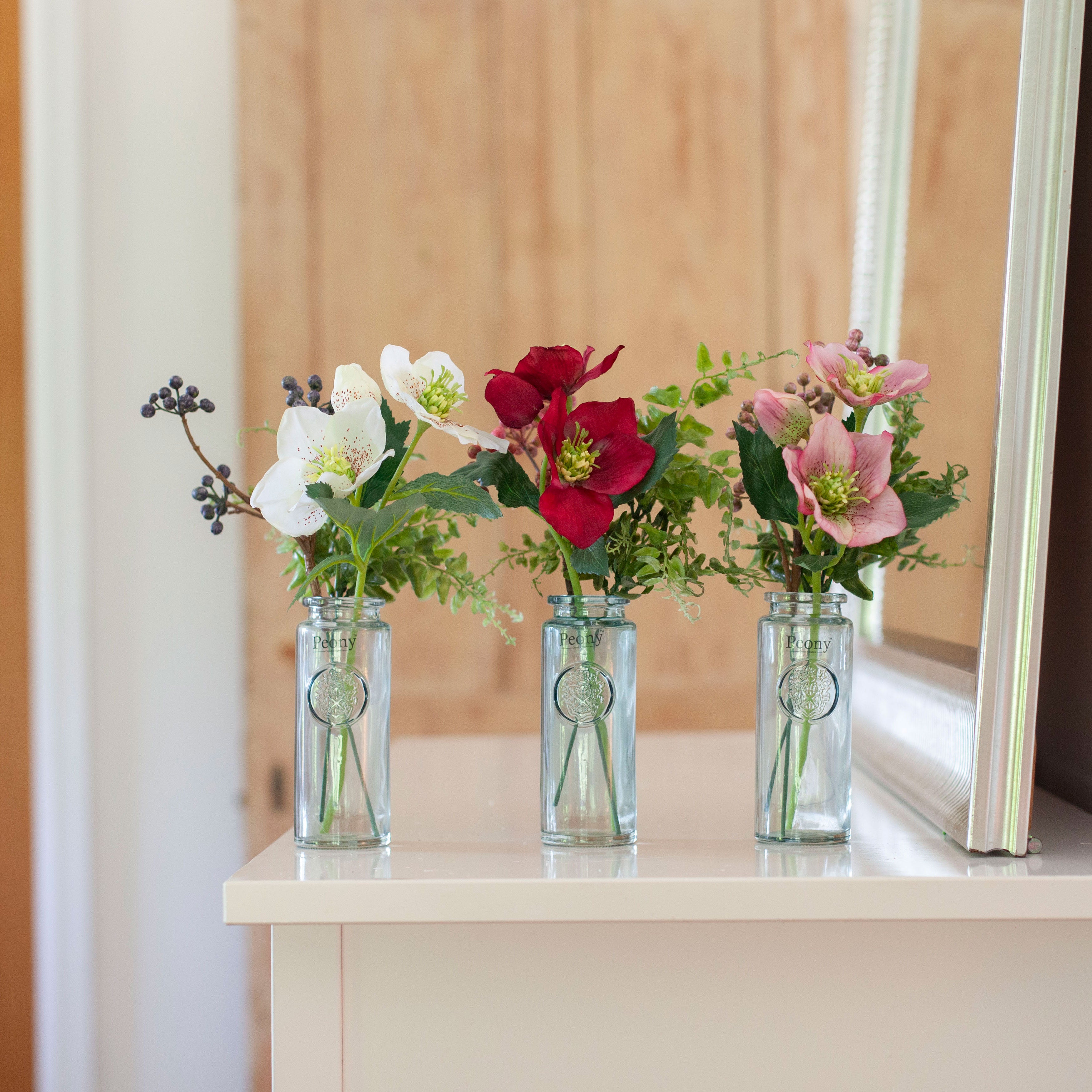 Set of three artificial hellebore and berry arrangements in white, burgundy and pink, arranged in individual glass bottle vases and styled on a console table.