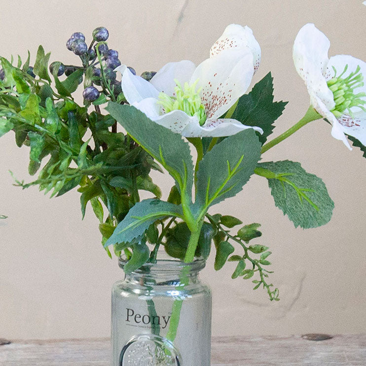 Close-up of an artificial white hellebore and berry arrangement with green foliage in a glass bottle vase