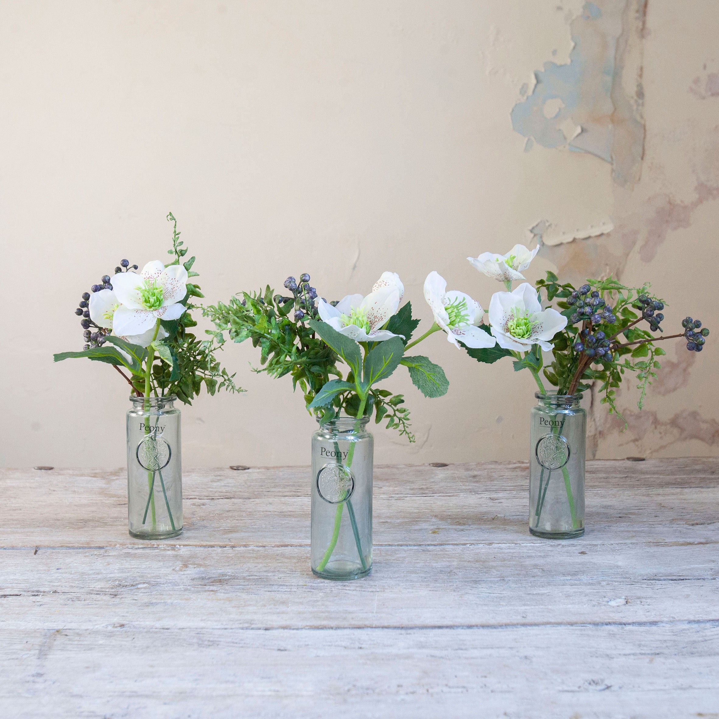 Set of three artificial white hellebore and berry arrangements with green foliage, arranged in individual glass bottle vases