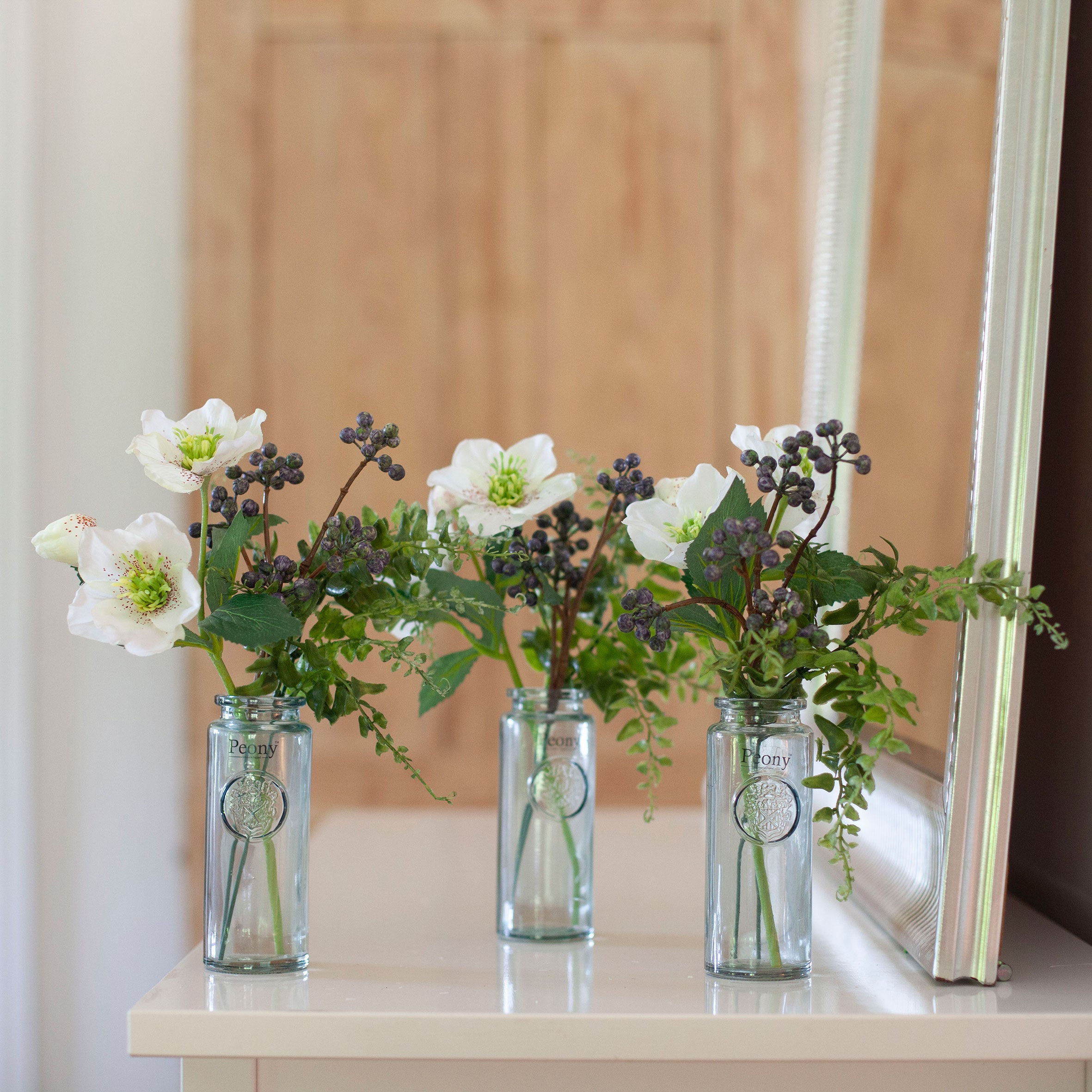 Set of three artificial hellebore and berry arrangements in  white, arranged in individual glass bottle vases and styled on a console table.