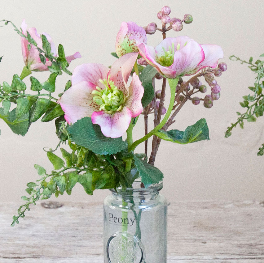 Close-up of an artificial pink hellebore and berry arrangement with green foliage in a glass bottle vase.