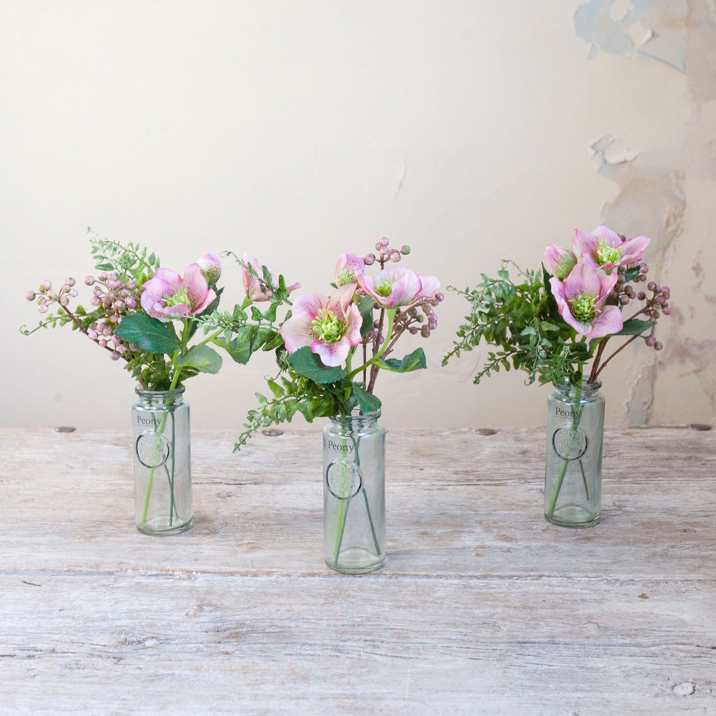 Set of three artificial pink hellebore and berry arrangements with green foliage in individual glass bottle vases.