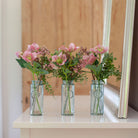 Set of three artificial hellebore and berry arrangements in  pink, arranged in individual glass bottle vases and styled on a console table.
